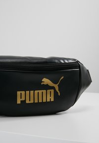 Banane noire de la marque Puma avec logo doré et fermeture éclair, présentée sur une surface blanche sur un fond gris uni.