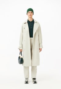 Ljusgrå dubbelknäppt trenchcoat, grön polo-t-shirt, beiga byxor, svarta sneakers och en rund grön handväska.