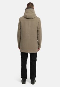 Lång, khakifärgad kappa med huva, tillverkad av mjukt tyg. Har en rak skärning och synliga sömmar längs ryggen. Kombinerad med svarta byxor.