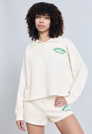 Donna con capelli ricci che indossa una polo a maniche lunghe color crema di Calvin Klein e pantaloncini abbinati, in piedi con una mano sui fianchi.