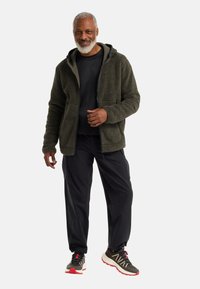 Olivgrüne Fleecejacke mit Reißverschluss, zwei Taschen, grauem Innenfutter, kombiniert mit schwarzen Cargohosen und Sportschuhen mit roten Akzenten.