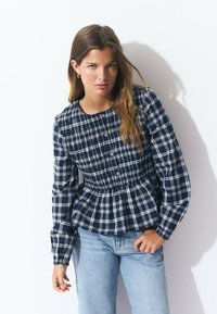 Checked navy and white long-sleeve top with a smocked waist and button details, paired with light blue jeans. Smooth cotton fabric.