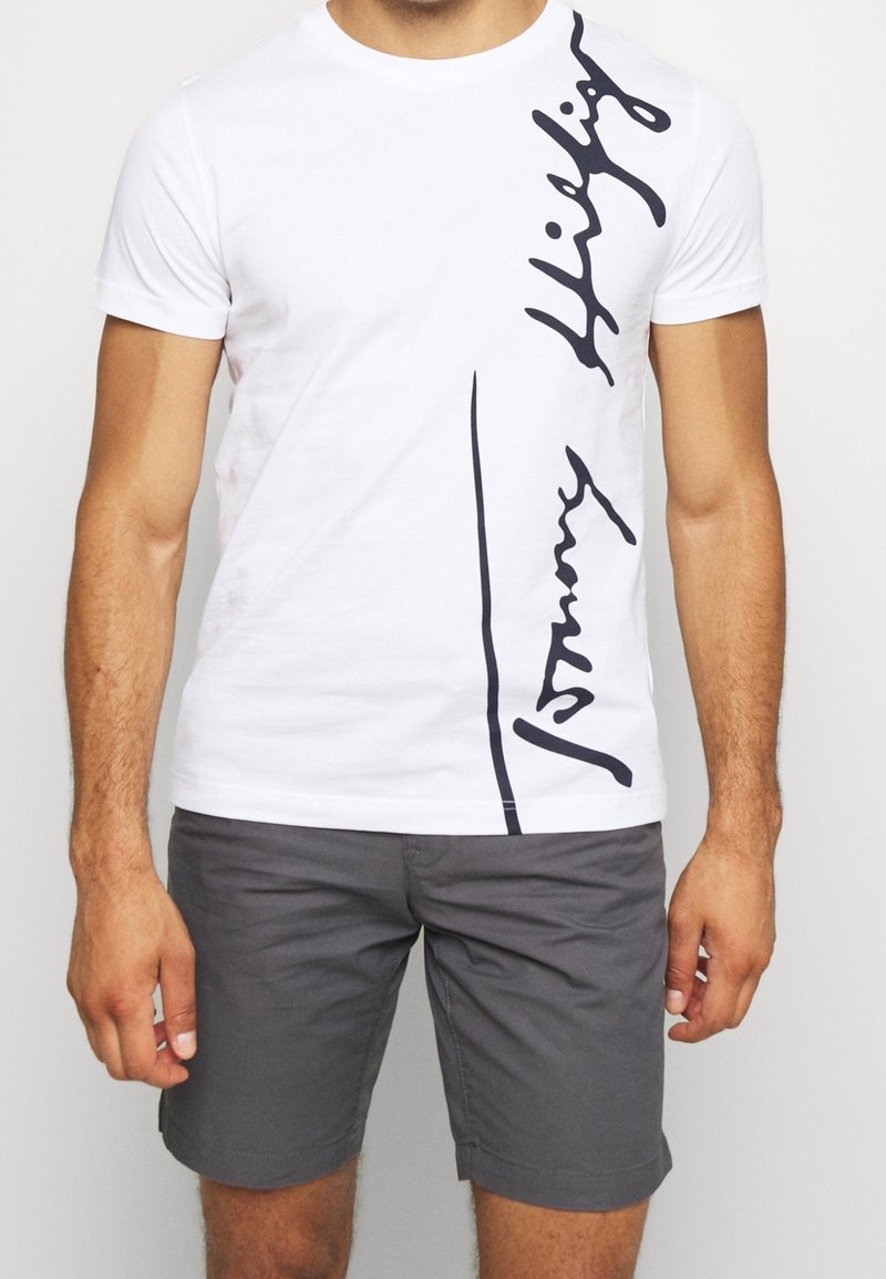 Camiseta blanca de manga corta con un logo de escritura negra diagonal en el lado izquierdo. Fabricada en algodón suave con un clásico escote de cuello redondo.