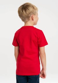 Blond boy facing away wearing a plain red short-sleeve T-shirt and dark blue pants against a white background.