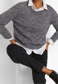 Personne portant un pull en maille gris par-dessus une chemise blanche à col, un pantalon noir et des baskets noires, assise avec une jambe croisée sur l'autre.