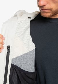 Leichte Jacke aus Mischgeweben: weiß mit einem strukturierten Muster, grauer Strukturpanel und dunkle Stoffakzente; enthält eine Reißverschlusstasche.