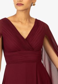 Nainen, joka käyttää viininpunaisia pleatattuja V-pääntiellä varustettuja mekkoja, joissa on läpikuultavat viittasleeveet ja kultaiset renkaakoru korvassa.