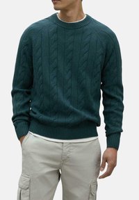 Suéter de punto verde oscuro con patrón de trenzado, cuello redondo acanalado y puños. Combinado con pantalones cargo beige claros.