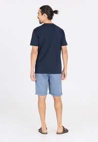 T-paita tummansinisestä puuvillasta, yhdistettynä vaaleansinseihin shortseihin, joissa on käänteet. Mustat liukuvasti jalkaan laitettavat sandaalit täydentävät asun.