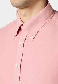 Chemise boutonnière rose clair avec un col classique, des boutons blancs et un petit logo brodé sur la poitrine. Texture lisse.