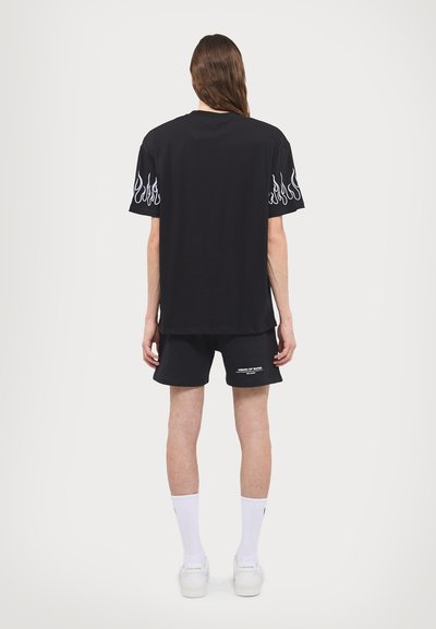 Must mustaht T-särk, millel on lühikesed varrukad ja leegiga graafika varrukatel, koos mustade lühikesega, millel on valge tekst. Mudel kannab valgeid sokke ja kingi.