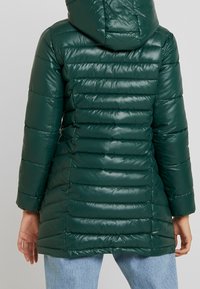 Chaqueta acolchada verde con capucha, acabado brillante, textura de acolchado vertical y corte a mediana longitud, combinada con jeans azul claro.