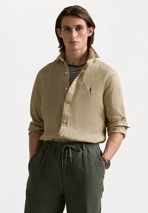 Camisa de lino beige con cuello abotonado, que presenta un pequeño logo en navy. Combinada con pantalones de tiro relajado verdes oliva con cordón.