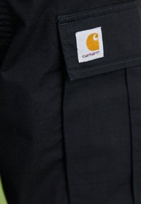 Gros plan sur une poche en tissu noir avec une étiquette carrée blanche affichant le logo jaune de Carhartt et le nom de la marque.