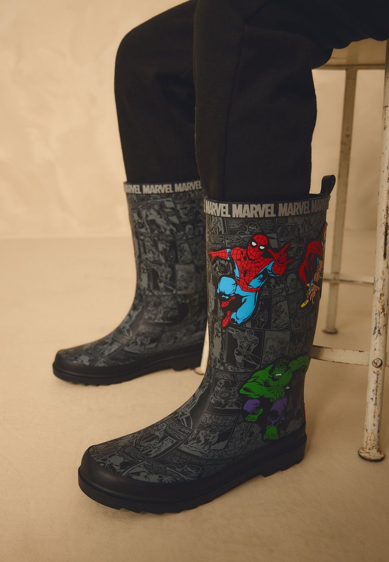 Bottes de pluie noires avec un design de bande dessinée Marvel et des images colorées de Spider-Man et Hulk portées par une personne assise sur un tabouret.