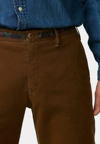 Braune Baumwollhose mit glatter Textur, ausgestattet mit einem gemusterten Gürtel und einem Knopfverschluss. Seitentasche in der Nähe des Bundes sichtbar.
