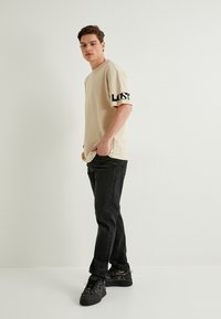 Beige bomulds t-shirt med "LOST" i sort skrift på ærmet, kombineret med tætsiddende sorte jeans og sorte sneakers.