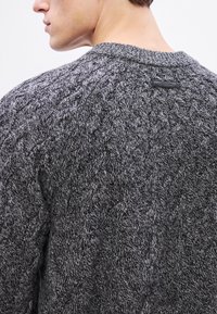 Biezs pelēks adīts džemperis ar teksturētu kabeļa rakstu, apaļu izgriezumu un nelielu melnu etiķeti uz muguras.