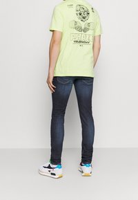 Camiseta de algodón verde neón con un diseño gráfico en la parte posterior, combinada con jeans de mezclilla oscura y zapatillas multicolores. Silueta ajustada.
