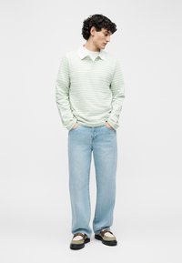 Nooremees lokkis juustega seisab ja vaatab alla, kandes heledat roheline triibulist pika varrukaga särki, helesiniseid teksaseid ja mokasiine.