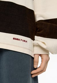 Kadar v bližini roke ob kremasto in temno rjavem prugastem puloverju z izvezenim napisom "BIMBA Y LOLA" blizu roba, v kombinaciji s temnimi hlačami.