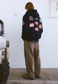 Tmavomodrá mikina s kapucňou s rôznymi nášivkami, v kombinácii s béžovými nákladnými nohavicami a hnedou obuvou, stojaca vedľa vozidla.