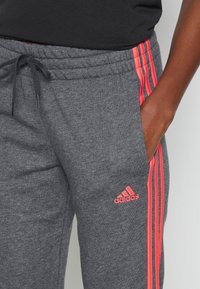 Harmaat collegehousut, joissa on joustava vyötärönauha ja kiristysnauha. Sivussa kolme vaaleanpunaista raitaa ja reiden kohdalla pieni vaaleanpunainen Adidas-logo.