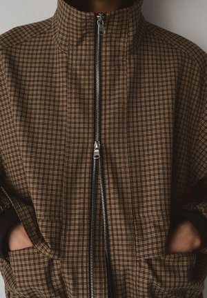 Persona con chaqueta a cuadros marrón y negra con puños negros, cremallera doble frontal parcialmente desabrochada, manos en los bolsillos de la chaqueta.