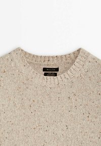 Beige neulepaita, jossa on teksturoitu pinta, ribatty pyöreä pääntie ja täplitetty yksityiskohta. Merkkilappu näkyvissä kauluksessa.