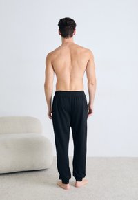 Schwarze Baumwoll-Jogginghose mit elastischem Bund, gefertigt für bequemen Tragekomfort, kombiniert mit einem unbedeckten Oberkörper. Weiche Textur, lässige Passform, Bündchen an den Knöcheln.