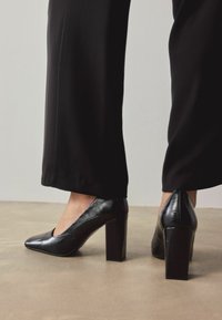 Czarne szpilki na wysokim obcasie z błyszczącym wykończeniem, ze spiczastym noskiem i masywnym, blokowym obcasem, z subtelnym teksturowanym wzorem.