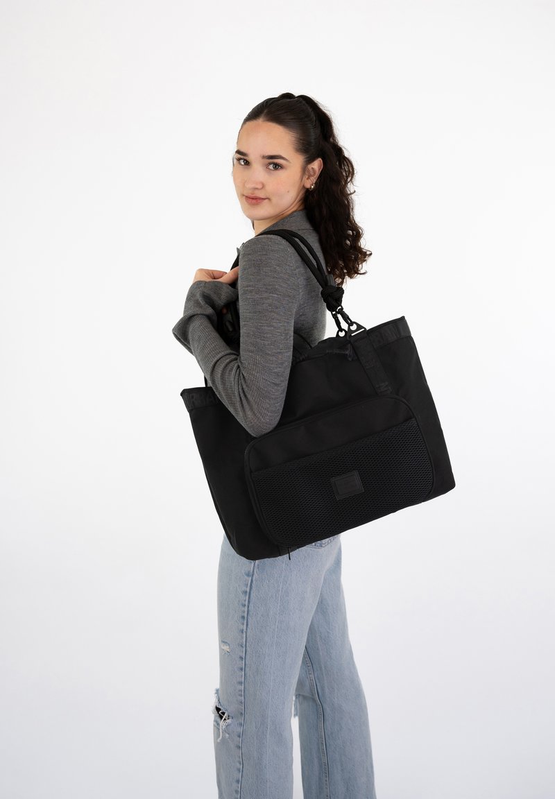 Black tote bag with a textured front pocket, dual handles, and adjustable strap. Fabric appears durable, featuring sleek lines and a minimalist design.