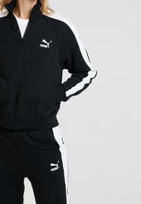 Svart zip-jacka med vita sidostråk, med hög krage och logotyp. Matchas med svarta byxor, båda i mjukt tyg.