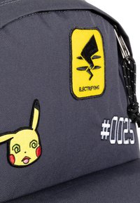Zaino in tessuto grigio con un patch giallo a forma di simbolo elettrico, un design di Pikachu con occhi a spirale e un'etichetta bianca "#0025".