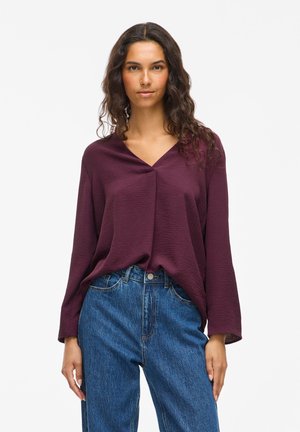 Kvinne med langt krøllete hår som har på seg en strukturert plommefarget langermet V-halsbluse som er stappet ned i høyt midjefetede blå jeans.