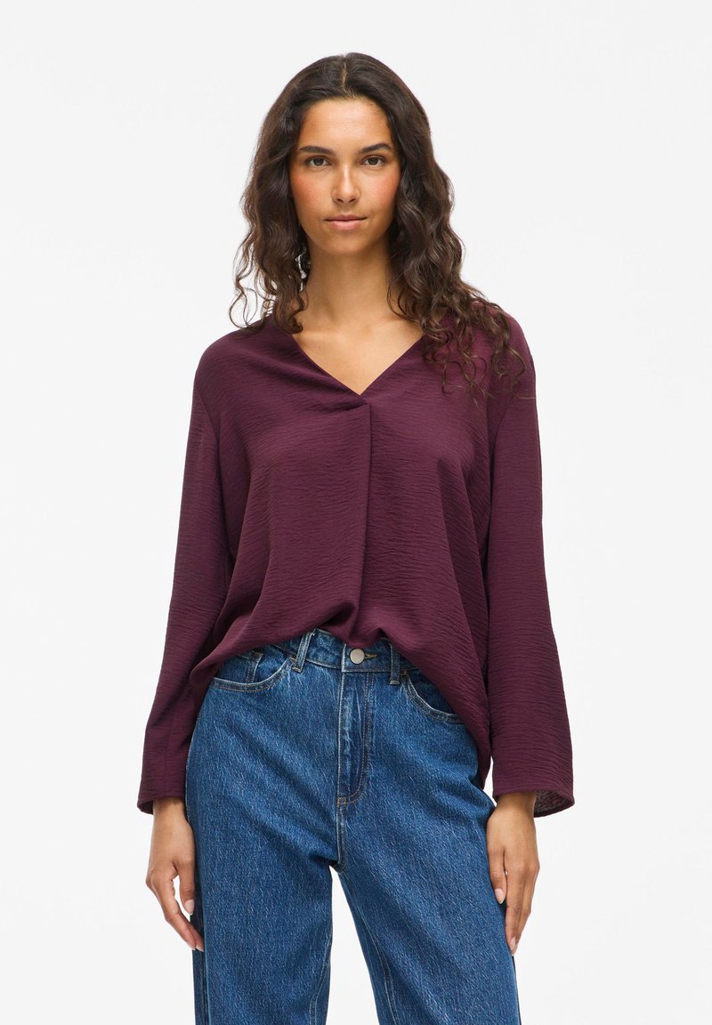 Frau mit langem, lockigem Haar, die eine strukturierte, pflaumenfarbene Bluse mit langen Ärmeln und V-Ausschnitt trägt, die in hochgeschnittene blaue Jeans gesteckt ist.