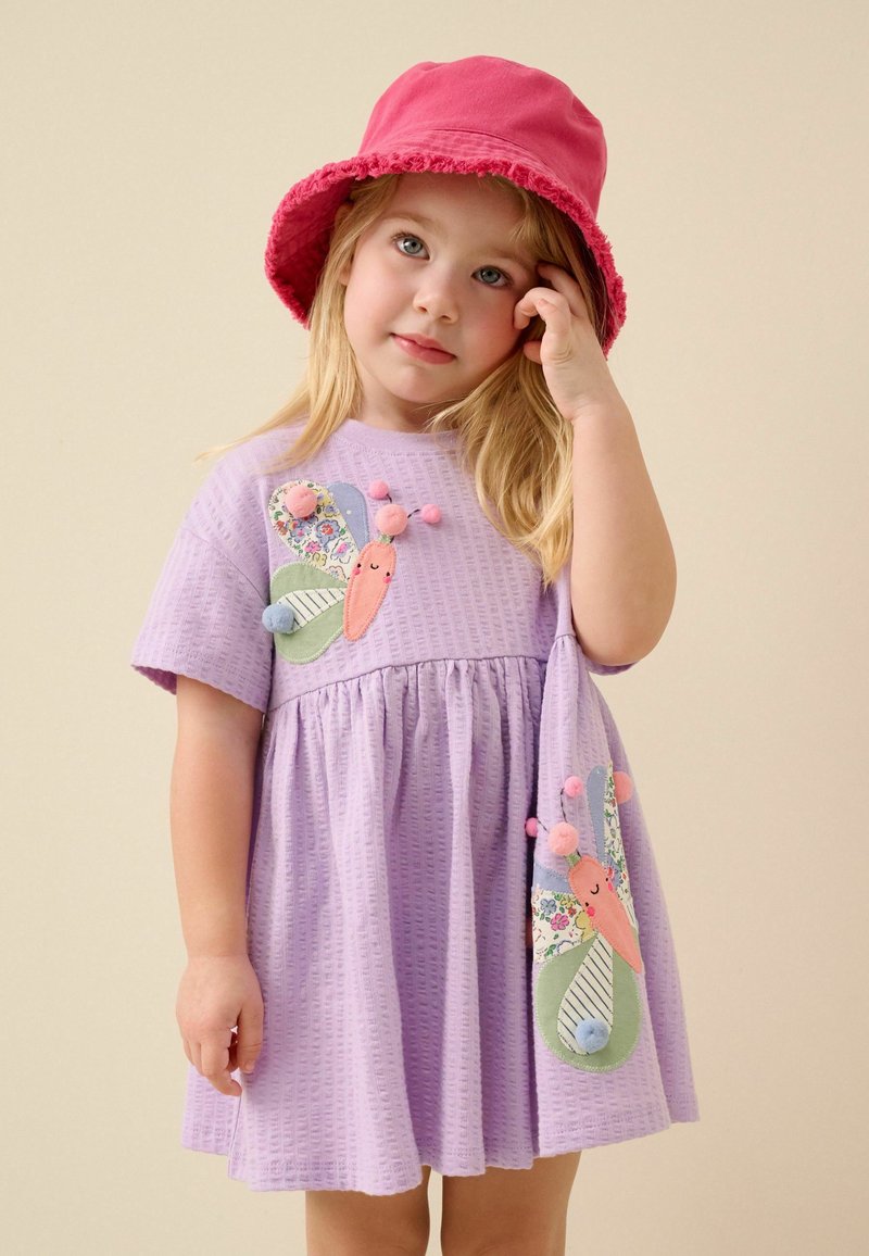 Jeune fille portant une robe violette avec des appliques papillons et un chapeau bob rose, touchant sa tête sur un fond beige neutre.