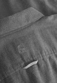 Grau gestreiftes Gewebe mit einer weichen Struktur, das ein genähtes &-Zeichen-Logo und einen umgeschlagenen Kragen aufweist. Der Stoff zeigt ein dezentes Webmuster.