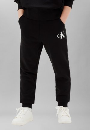 Person trägt schwarze Calvin Klein Jogginghosen mit Taschen und weiße Sneaker, steht mit den Händen in den Taschen vor einem schlichten Hintergrund.