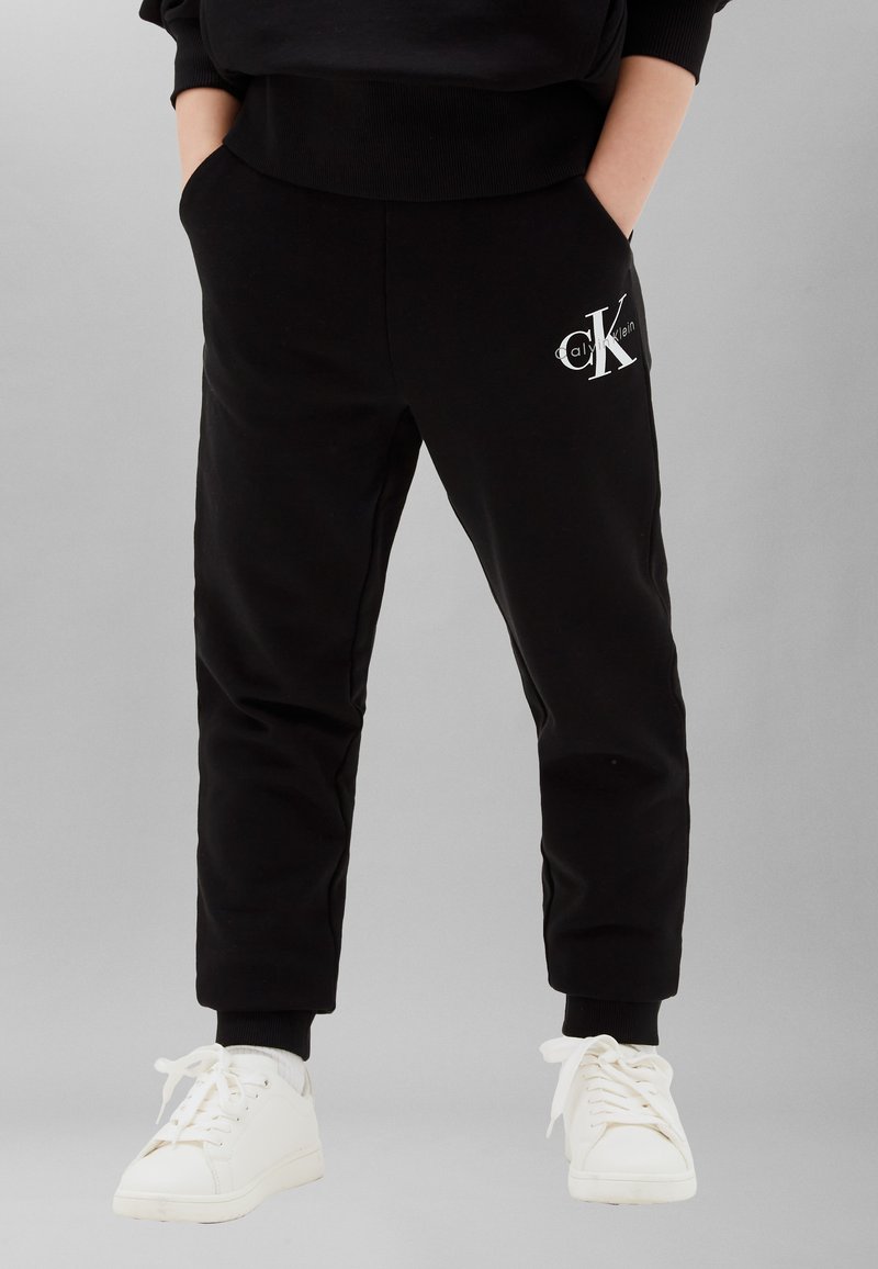 Άτομο που φοράει μαύρα joggers Calvin Klein με τσέπες και λευκά αθλητικά παπούτσια, στέκεται με τα χέρια στις τσέπες μπροστά σε ένα απλό φόντο.