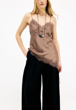 Ženska v rjavi satenasti srajčki z čipkastim robom, črne široke hlače in črno-bela obesek ogrlica na belem ozadju.