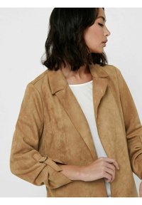 Nainen, jolla on olkapitkät tummat hiukset, pukeutuneena vaaleanruskeaan nahkatakkiin valkoisen topin päällä, katsoo sivulle yksinkertaista taustaa vasten.