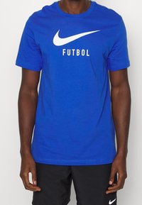Modré bavlněné tričko s velkým bílým logem Nike a slovem "FUTBOL" tučnými černými písmeny, s krátkým rukávem a kulatým výstřihem.