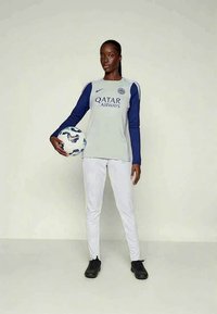 Nainen jalkapalloilija harmaan ja laivansiniset pelipaidat päällä, pitäen palloa kädessään, valkoisissa housuissa ja mustissa nappulakengissä, seisoo yksinkertaista taustaa vasten.