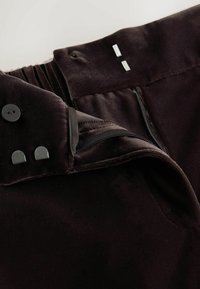 Nahaufnahme eines schwarzen Samtkleidungsstücks mit Reißverschluss, Knöpfen und Metallakzenten. Weicher Stoff mit gefältelten Details an der Taille.