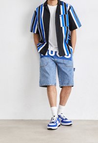 Man in een blauw, zwart en wit gestreept overhemd met korte mouwen, een wit t-shirt, denim shorts met een opvallend blauw logo, witte sokken en blauwe sneakers.