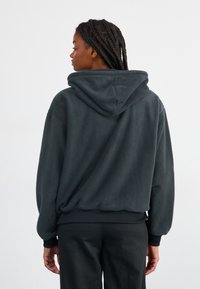 Tmavě šedá mikina s kapucí, zobrazená ze zadní strany, s jemnou texturou, uvolněným střihem a žebrovanými manžetami na pase a rukávech.