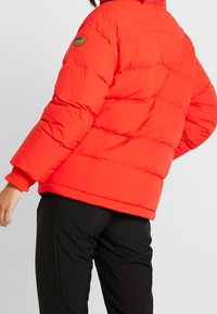 Czerwona kurtka puchowa z pikowanym wzorem, zaokrąglonym kołnierzem i elastycznymi mankietami. Posiada małą naszywkę z logo na górnej lewej rękawie.