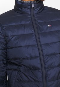 Veste matelassée bleu marine avec des coutures horizontales, fermeture éclair sur le devant, petit logo de la drapeau Tommy Hilfiger sur la poitrine et bordure de col brandée.
