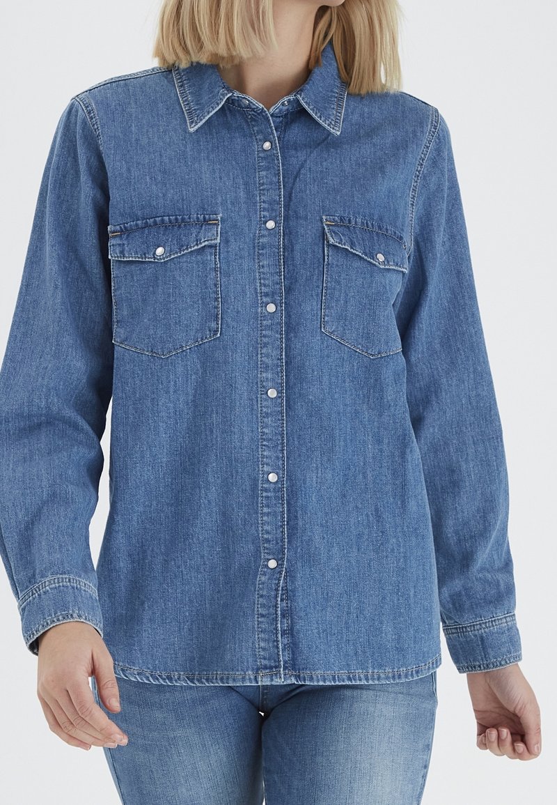 Femme portant une chemise en jean bleue à manches longues boutonnée avec deux poches poitrine et un jean bleu assorti, debout devant un fond uni.